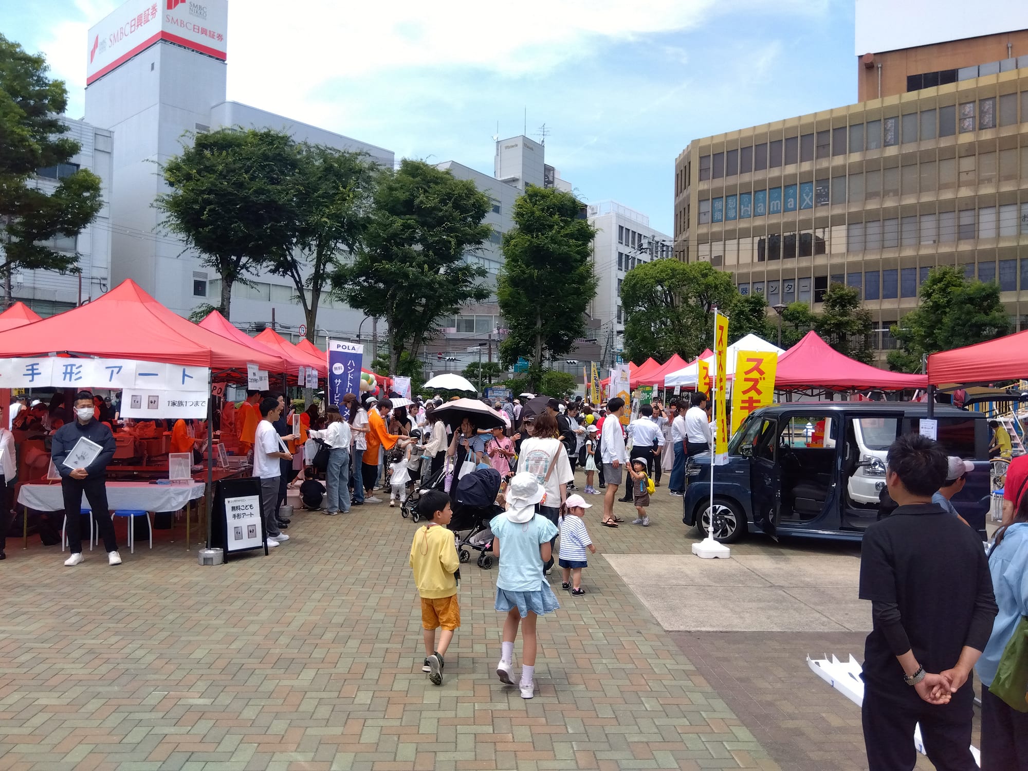 屋外のイベント会場。赤いテントのブースが並び、家族連れの来場者が行き交っている