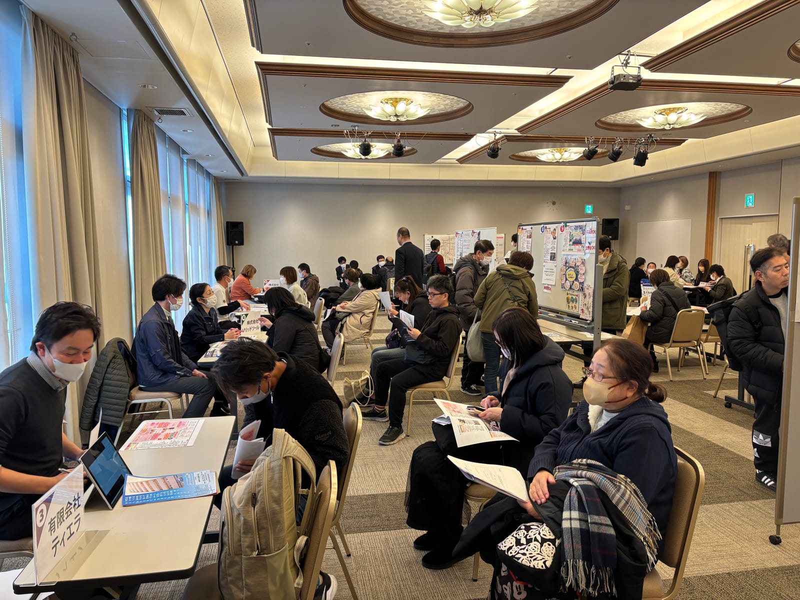 企業のテーブルで採用担当者と来場者が向かい合って話し、順番を待つ来場者が資料を読んでいる